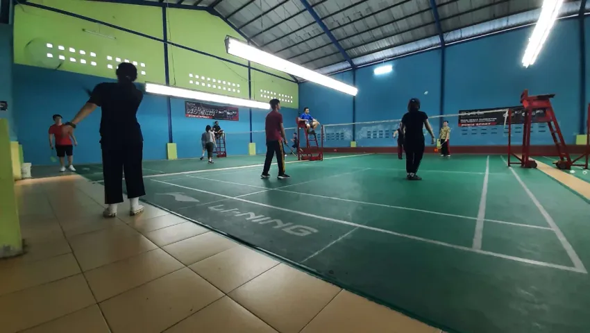 Wide-angle view of an indoor badminton court with players in a green-floored hall with a blue wall.