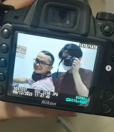 A camera's back screen showing a photo of two people smiling, one holding a camera up to their face. The screen shows the file number, the date 09/12/2023 and the image size.
