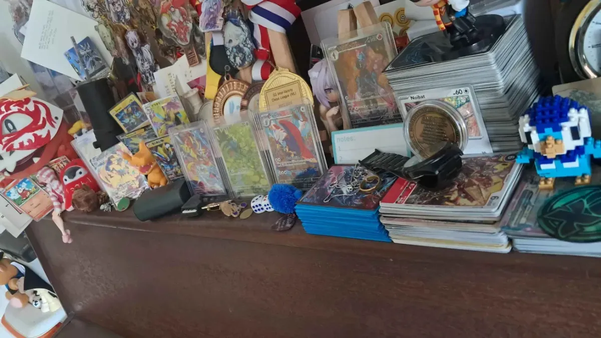 A wooden shelf covered in stacks of sleeved trading cards, cards in hard cases, medals, small figures and a Piplup pixel-brick figure.