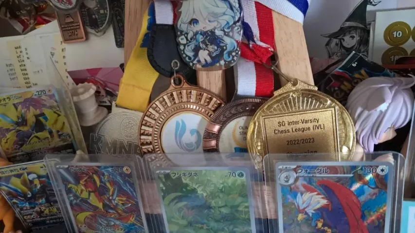 A pile of medals in front of framed Pokemon cards, including a gold medal for the SG Inter-Varsity Chess League (IVL) 2022/2023.